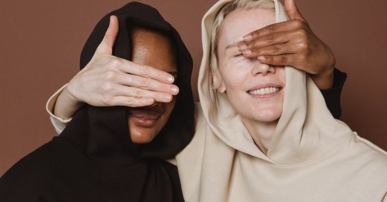 Content Creation Trends - Content diverse female models in casual hoodies covering eyes of each other with hands while standing on brown background in studio