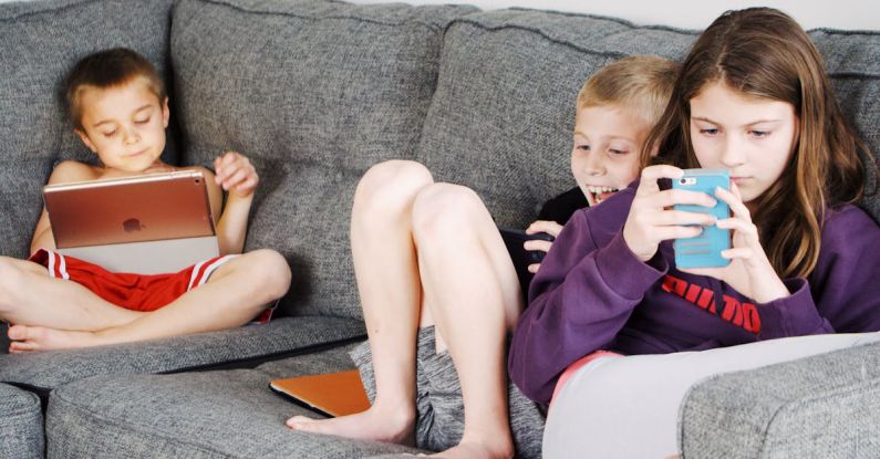 Smartphone Games - Positive barefoot children in casual wear resting together on cozy couch and browsing tablets and smartphones